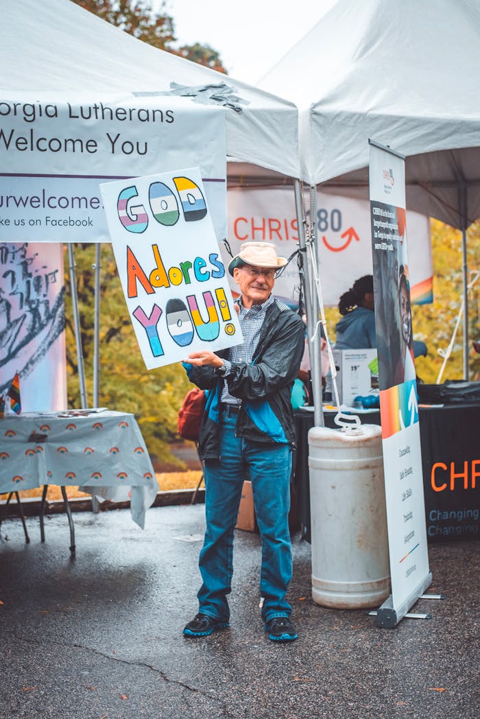 our-story-01 Elderly man smiling while holding a vibrant religious sign at an outdoor community event.